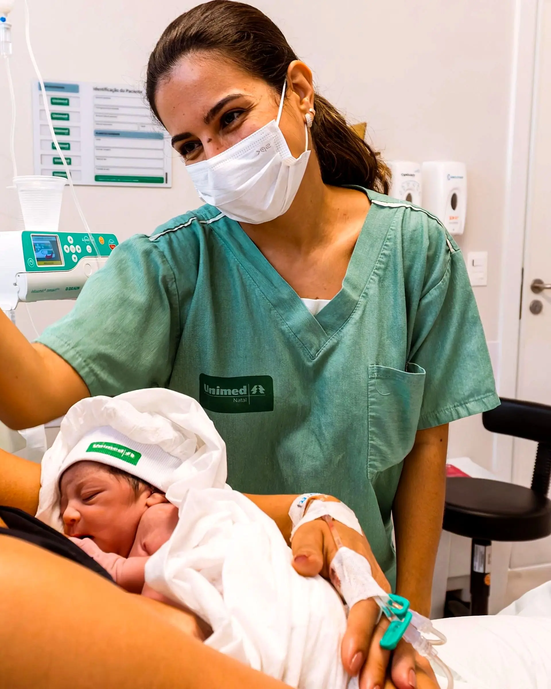 Gabriela, de pijama cirúrgico verde, sorrindo para uma mãe que segura o recém-nascido no quarto do hospital