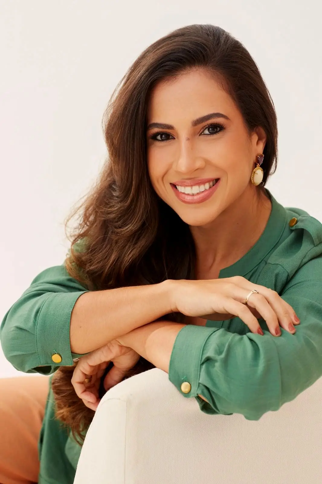 Gabriela de camisa verde, sorrindo, com os braços apoiados no encosto de uma poltrona